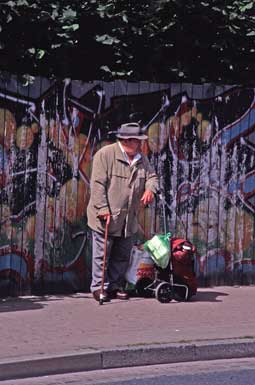 Mann mit Einkaufswagen
                und Tueten vor Graffiti-Holzzaun